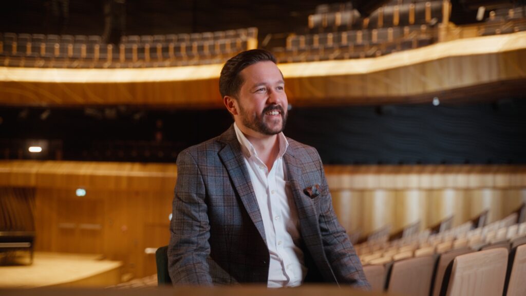 Cavatina Philharmonic Orchestra: A smiling Jacek Obstarczyk in a plaid blazer and white shirt sits in an empty auditorium, looking to the side with a cheerful expression.