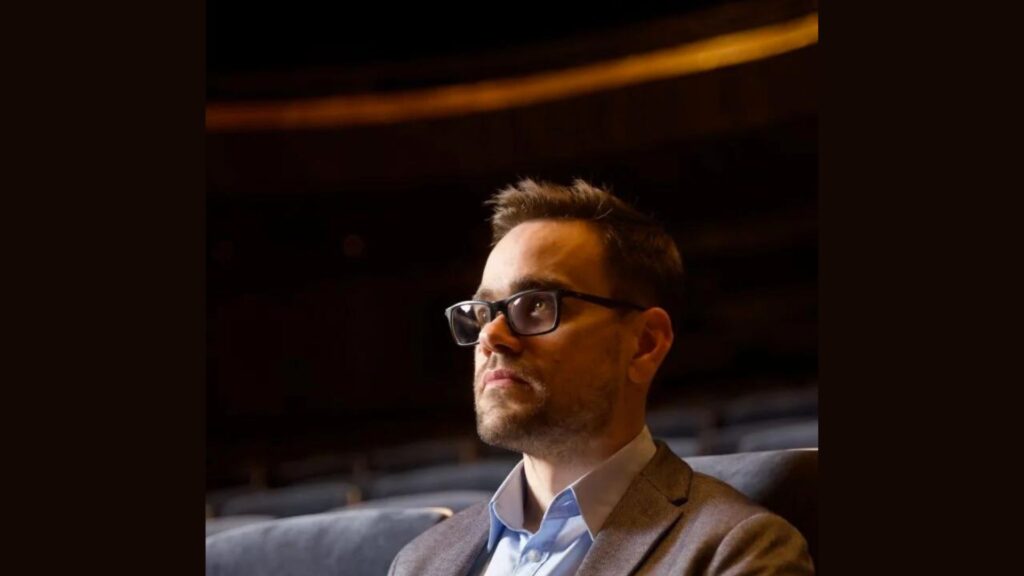 Cavatina Philharmonic Orchestra: Jacek Stolarczyk, with glasses and a serious expression, sits alone in an auditorium, gazing thoughtfully ahead.