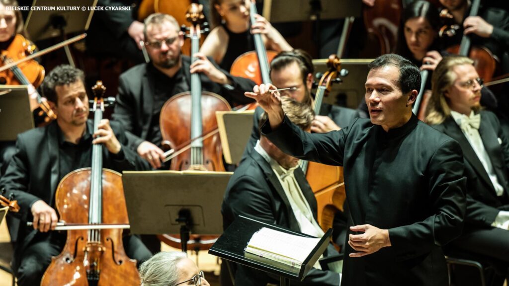 Cavatina Philharmonic Orchestra: The conductor leads the orchestra during a performance, with musicians focused on their instruments in the background.