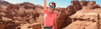 Autoimmune Diseases: A smiling Maria Mazurek in a pink shirt and sunglasses raises her fist triumphantly while standing in front of a scenic desert canyon under a clear blue sky.