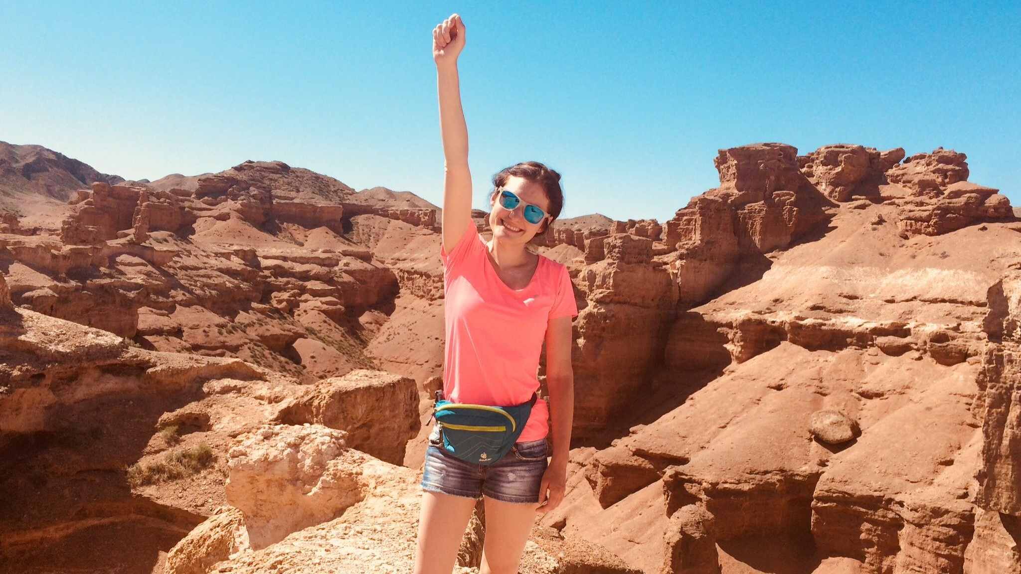 Autoimmune Diseases: A smiling Maria Mazurek in a pink shirt and sunglasses raises her fist triumphantly while standing in front of a scenic desert canyon under a clear blue sky.
