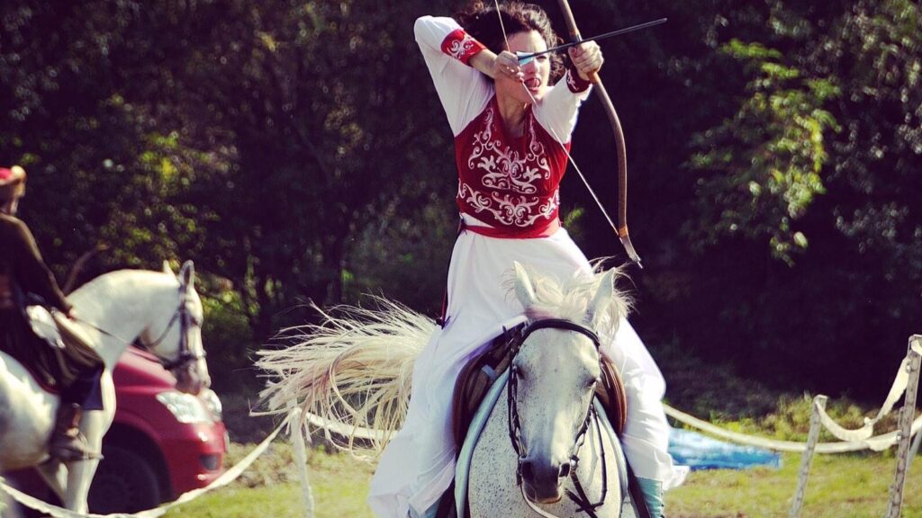 Autoimmune Diseases: Maria Mazurek in traditional attire skillfully shoots an arrow while riding a white horse during an outdoor archery event.