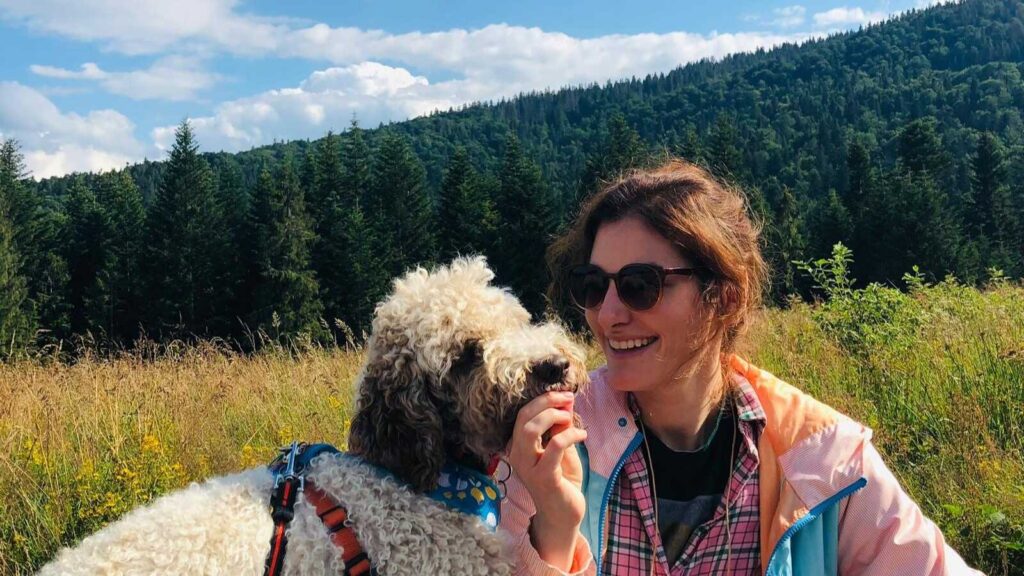 Autoimmune Diseases: Maria Mazurek in sunglasses smiles while interacting with a curly-haired dog in a scenic mountain meadow.