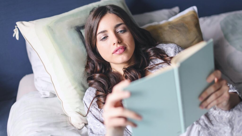 Biography and Truth: A young woman with long brown hair lies on a bed, comfortably reading a book