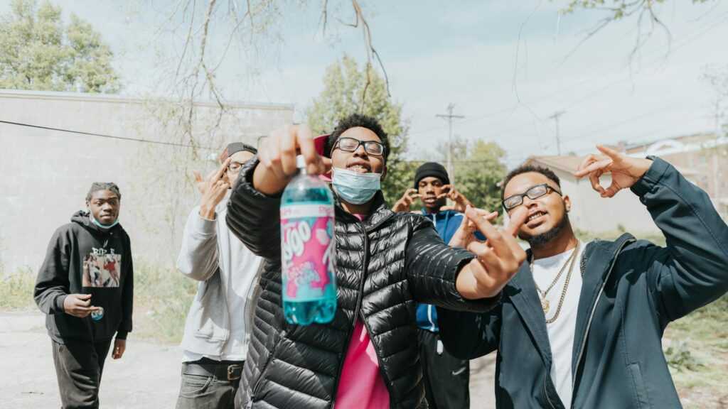 Crime in Sweden: A group of young men pose together outdoors, some making hand gestures, with one holding a bottle of soda and wearing a face mask.