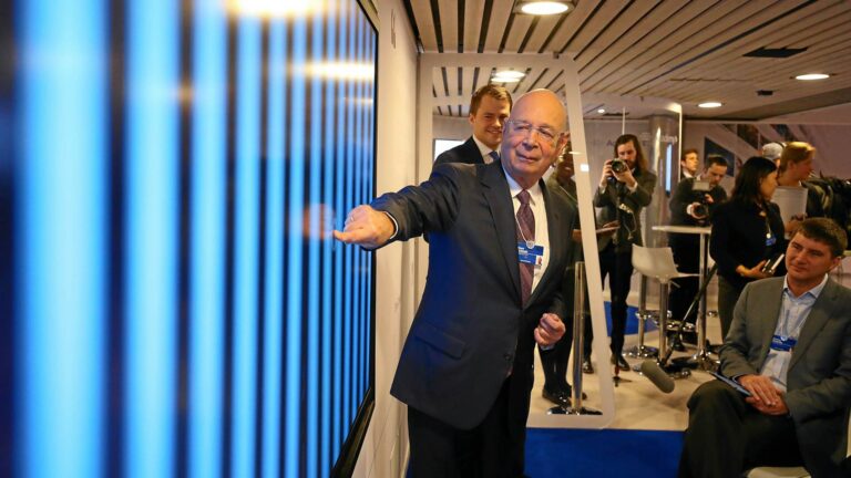 An elderly man in a suit touching a large TV screen with his finger