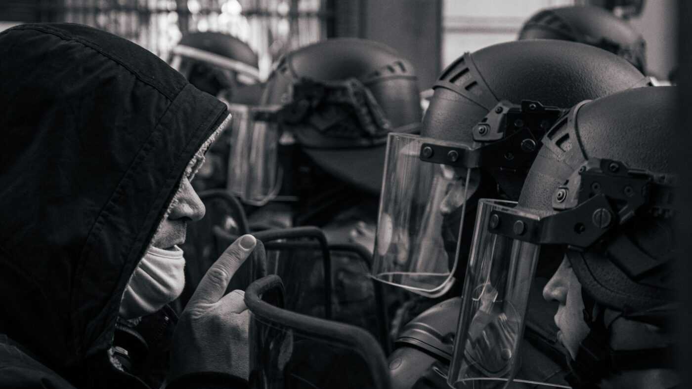 Immigrants in France: A protester in a hooded jacket confronts a line of French riot police officers wearing helmets and face shields.