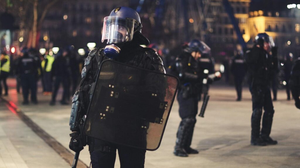 Immigrants in France: A French riot police officer in full gear, holding a shield, stands in a public square during a protest at night in Lyon.