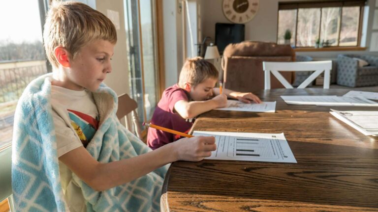 PISA Test: Two young boys sit at a table working on school assignments, with one wrapped in a blue blanket.