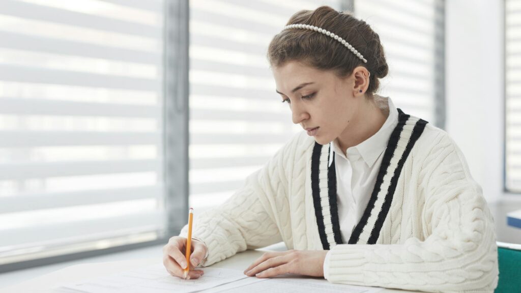 PISA test: A young woman wearing a white sweater and a pearl headband concentrates on writing at a desk.