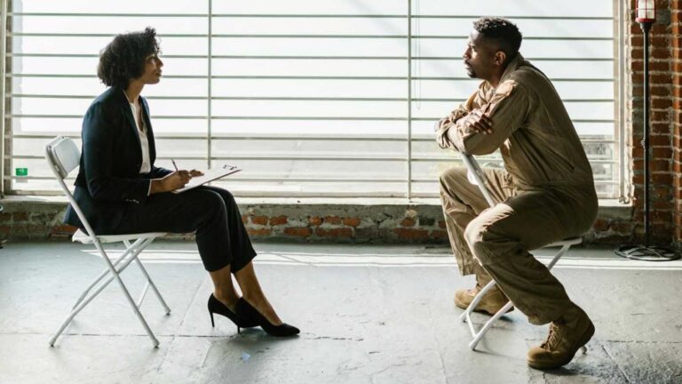 Pseudoscience and Psychotherapy: A woman in a professional suit and high heels sits on a folding chair with a clipboard, facing a man in a jumpsuit sitting on another folding chair. They are in a spacious, industrial room with large windows and exposed brick walls. The scene appears to depict a serious conversation or interview.