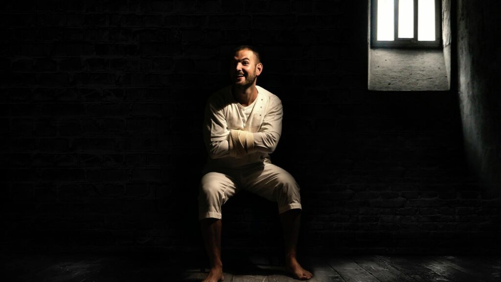 Psychopaths and Crime: a smiling and deranged man in a straitjacket in a dark cell with a barred window behind him