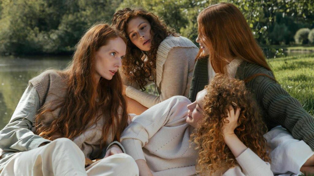 The Beauty Myth: Four women with long, wavy hair sit and lie on the grass near a serene pond in a peaceful outdoor setting