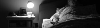 What do we regret: A grayscale picture of an elderly women lying on the bed, the room covered in shadows, a nearby lamp is the only source of light.