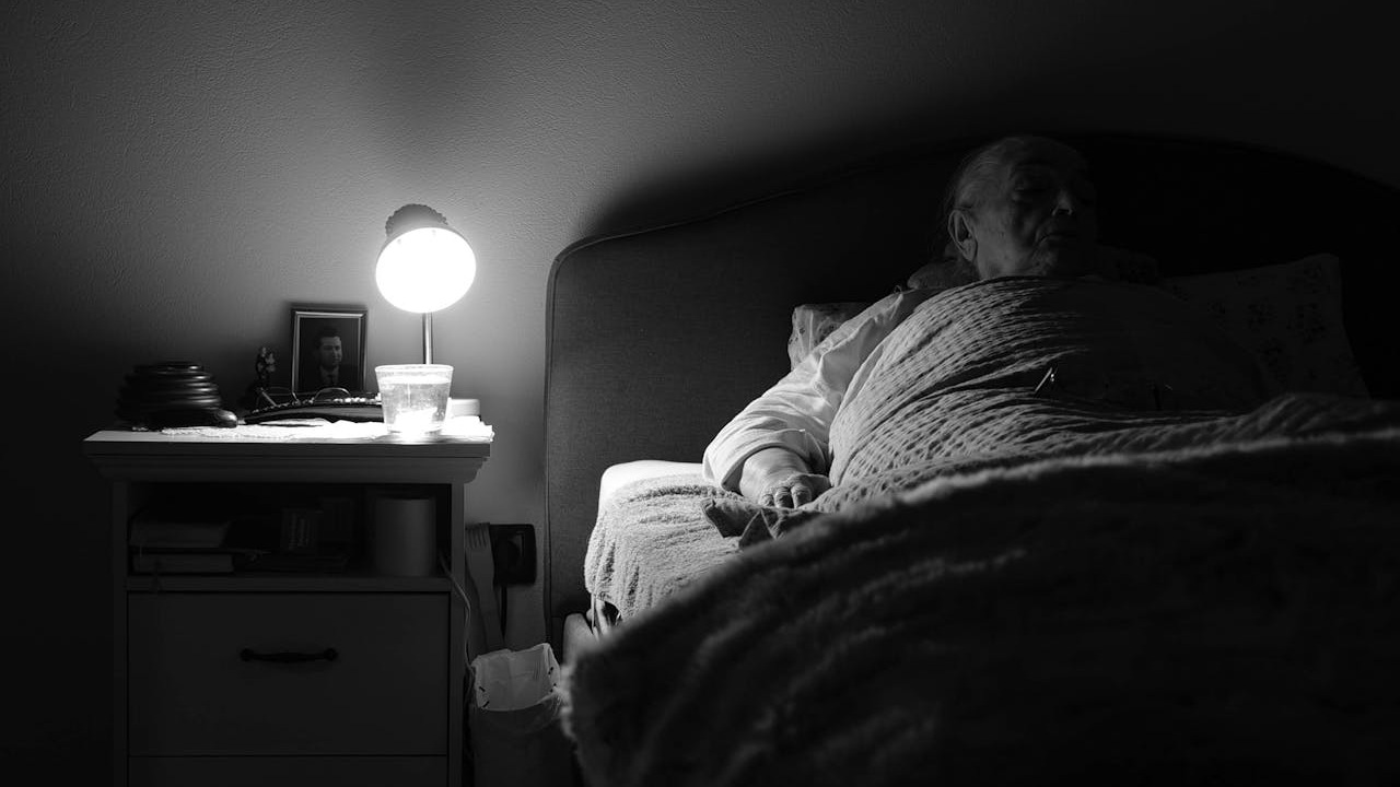 What do we regret: A grayscale picture of an elderly women lying on the bed, the room covered in shadows, a nearby lamp is the only source of light.