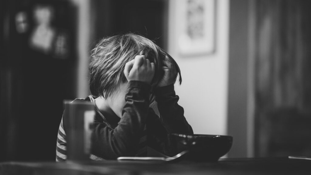 Burnout Kids: A black and white image of a child sitting at a table with their head in their hands, appearing distressed.