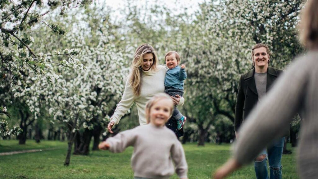 Family History: A family enjoying a day outdoors, with a mother holding a toddler and a young child running joyfully in a blooming orchard.