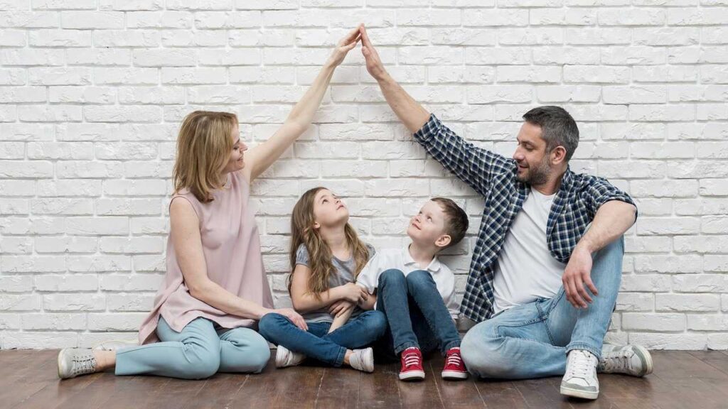 Family History: A family of four sitting on the floor against a white brick wall, with the parents forming an arch with their hands over the heads of their two children.