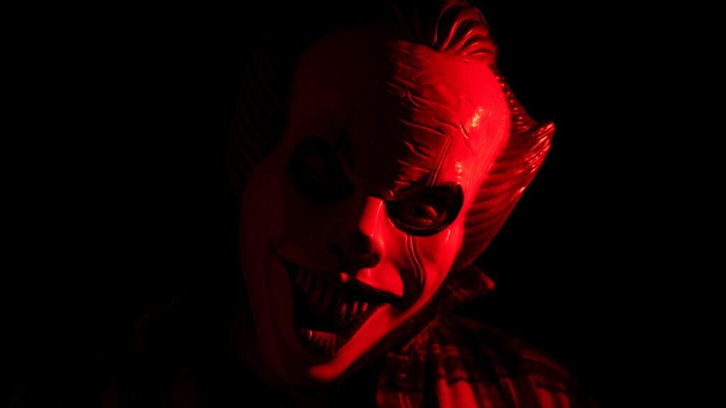 Fear and the Brain: A close-up of a menacing clown mask illuminated in red light, with a dark background creating a sinister atmosphere.