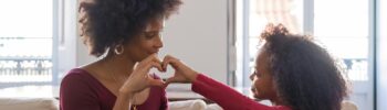 Feminism and Motherhood: A mother and daughter sit on a couch, forming a heart shape with their hands, sharing a tender moment together.