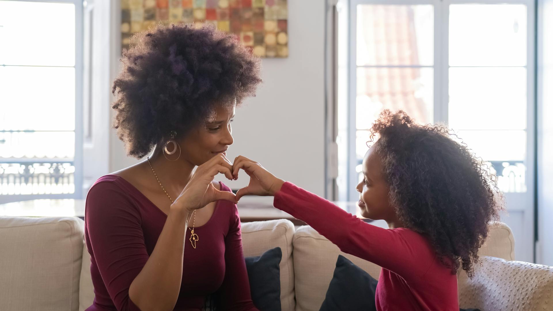 Feminism and Motherhood: A mother and daughter sit on a couch, forming a heart shape with their hands, sharing a tender moment together.