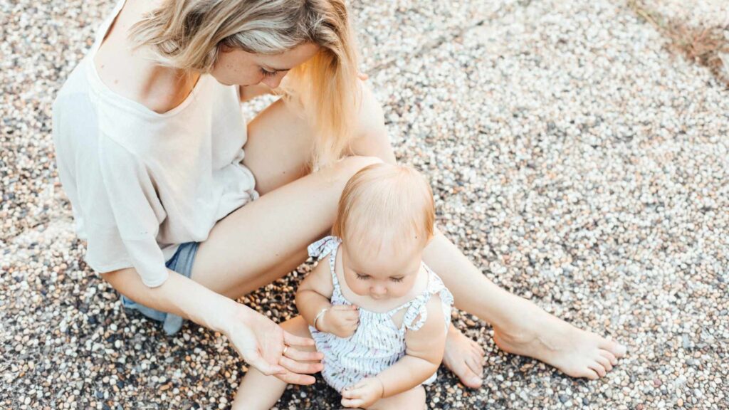 Feminism and Motherhood: A mother sits on the ground with her baby, gently interacting as they enjoy a quiet, sunny moment together.