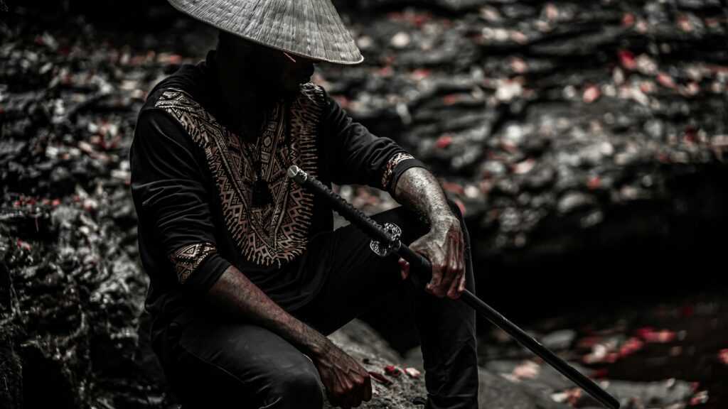 Honor Code: A person wearing a traditional conical hat, sitting in a dark, forested area with a sword resting on their lap.