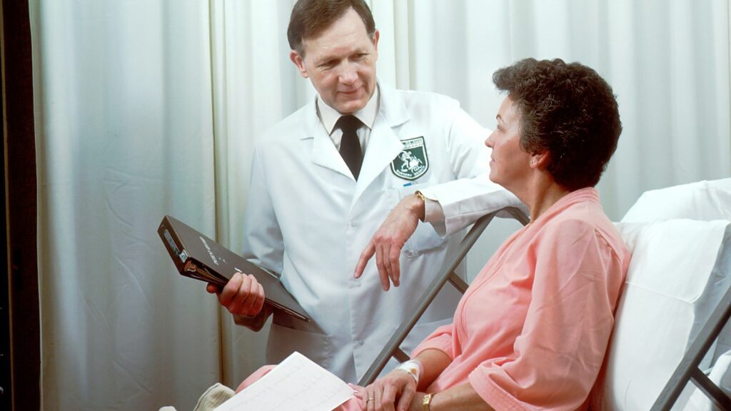 Medical Breakthroughs: A doctor in a white coat, holding a medical chart and showing concern, speaks with a patient in a hospital gown.