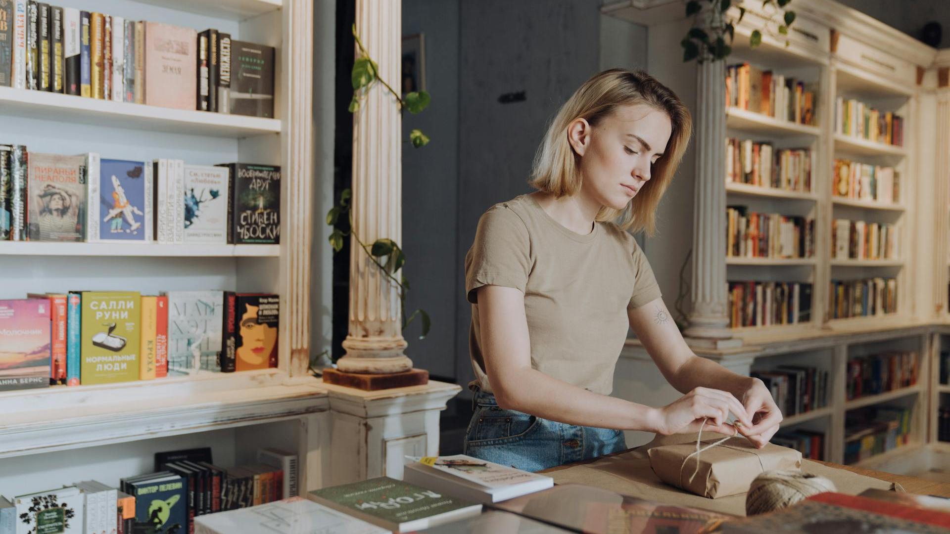 Modern Bestsellers: A woman carefully wraps a book in brown paper in a cozy bookstore filled with books on wooden shelves.