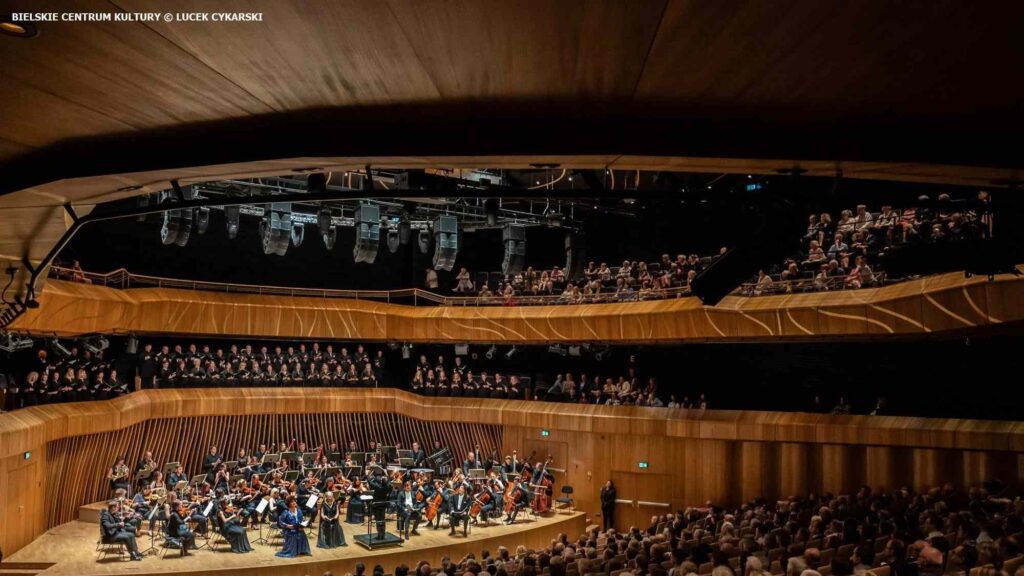 Private Orchestra in Poland: An orchestra and choir perform on stage in a large, modern concert hall filled with an attentive audience.