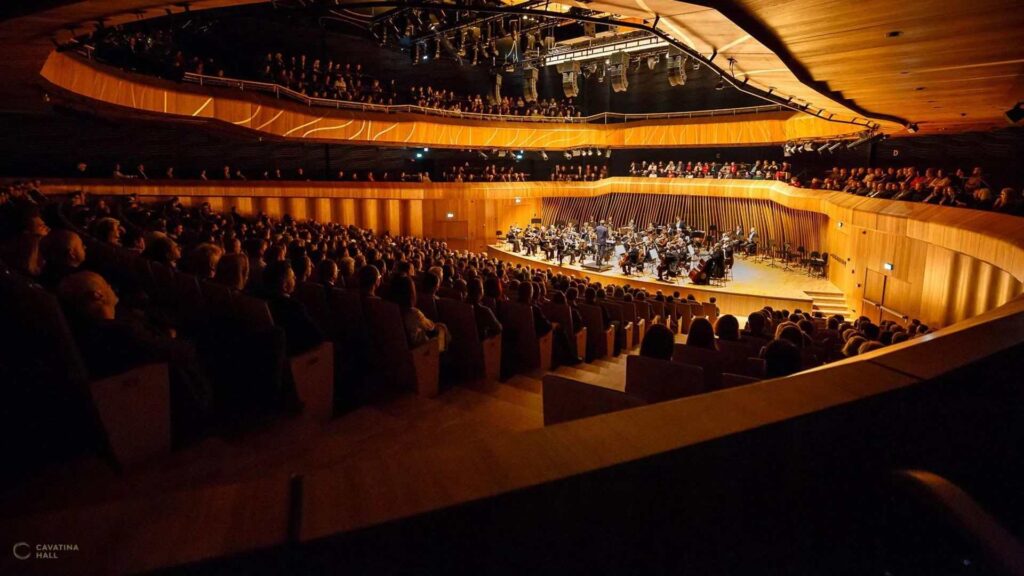 Private Orchestra in Poland: An orchestra performs on stage in a packed, warmly lit concert hall with audience members seated throughout.