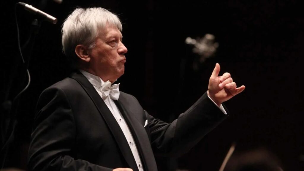 Private Orchestra in Poland: An older conductor with gray hair and a white bow tie conducts the orchestra with a focused expression and precise hand movements.