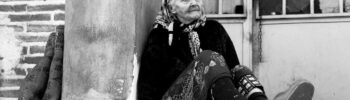 Senior Loneliness: an elderly woman sitting on the ground next to an old house.