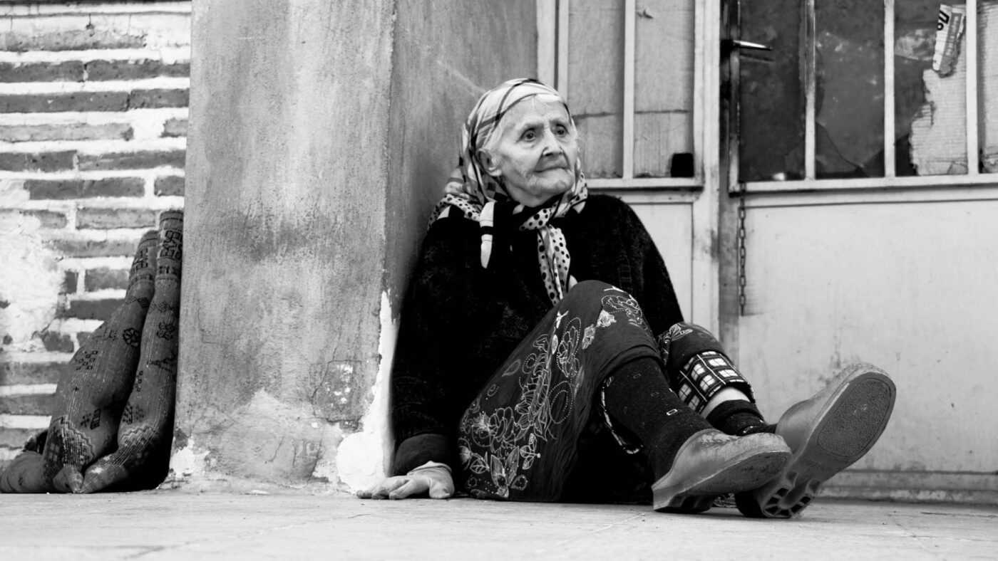 Senior Loneliness: an elderly woman sitting on the ground next to an old house.