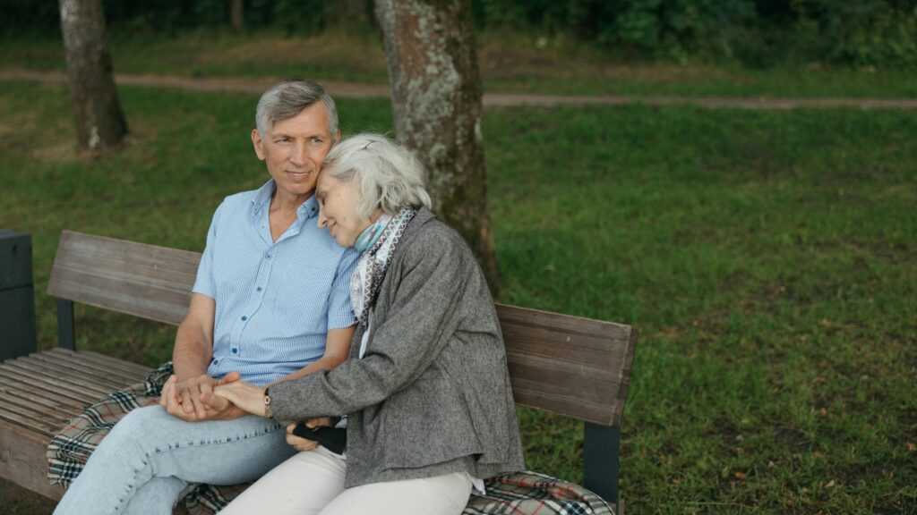 Senior Loneliness: a gray-haired couple on a bench in a park. They are holding hands