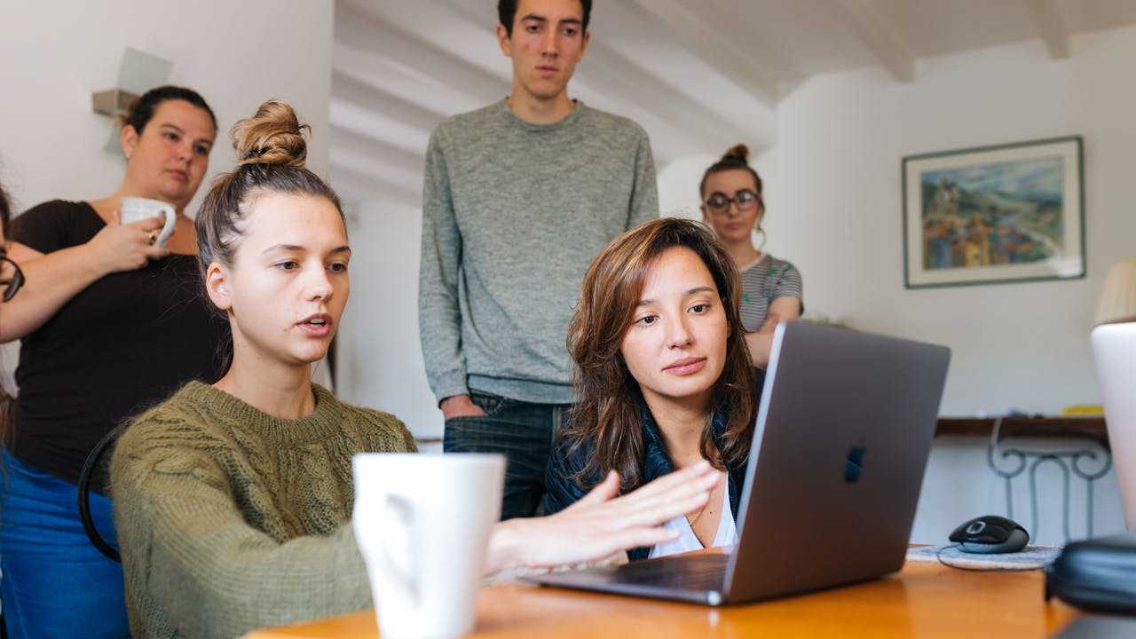 Working in a Group: A woman in front of a laptop explains something while the group of people around her watch the screen intensively