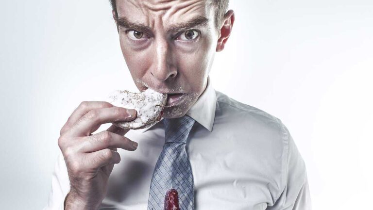 Cortisol-lowering foods, Photo: RyanMcGuire/pixabay. A stressed man in a stained shirt eats a donut, looking directly at the camera with a tense expression.