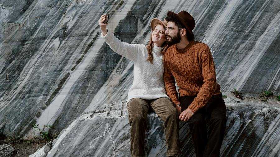 The compulsion to travel: a woman and a man in sweaters and hats taking a selfie in front of a rock formation. Fot. cottonbro studio / Pexels