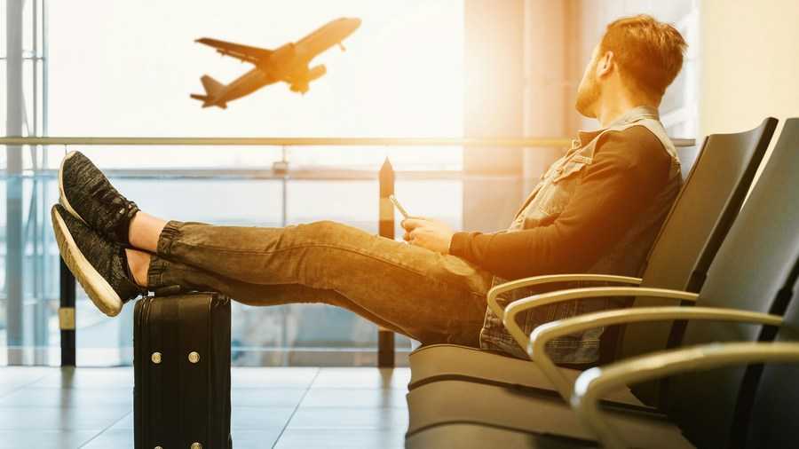 The compulsion to travel: a man sits in an airport waiting room with his feet resting on a suitcase, looking at a plane outside the window. Fot. JESHOOTS.COM / Pexels