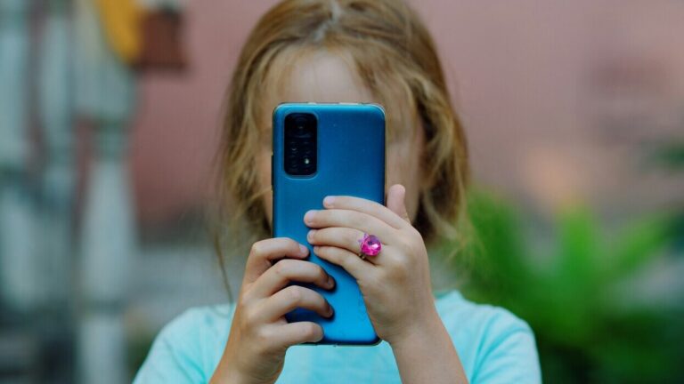 A child holding a smartphone in front of their face, using the phone in an urban space.
