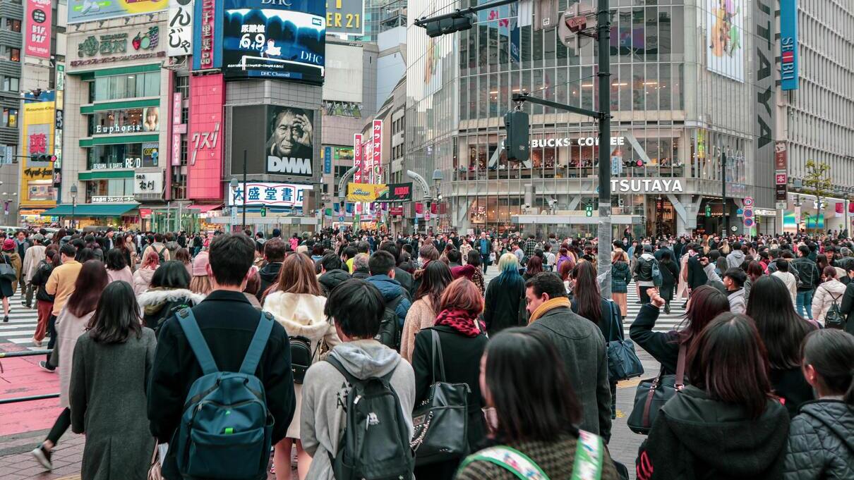 A crowd of people at a busy intersection in a large city, surrounded by noise, advertisements, and intense traffic, illustrating the impact of urban stress.