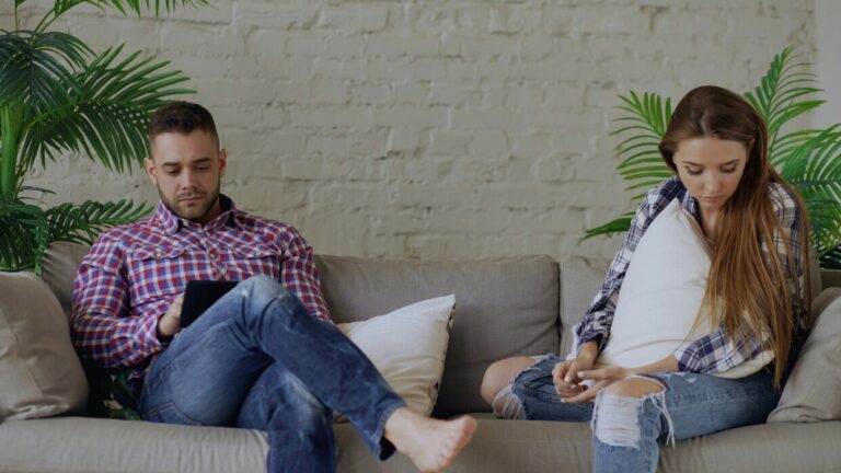 Two adults sit side-by-side on a sofa, each preoccupied with something else—a picture of a relationship that looks like a committed bond but functions like a no-strings-attached arrangement. This shot perfectly illustrates a situationship: physically together, yet emotionally apart.