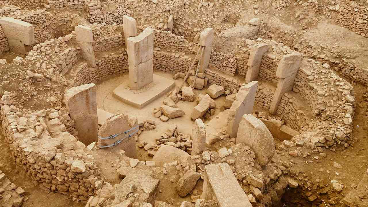 An aerial view of an archaeological excavation site showing a large, rectangular mass grave. The image displays distinct stratigraphic layers of soil and sediment, with marked sections where researchers identified human remains from the 14th century.