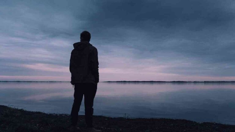 A figure stands alone by calm waters at twilight. In the apparent silence, we see an experience many of us know all too well—loneliness in a crowd, even when the world around us seems full of possibilities.