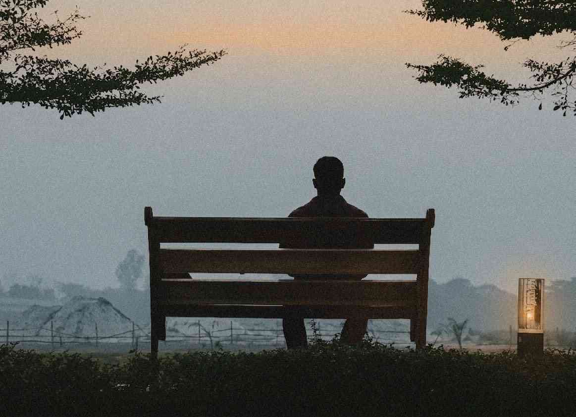A lonely man sits on a bench in a park at dusk, surrounded by silence and deep in thought. He reflects on life, fear, and hope.