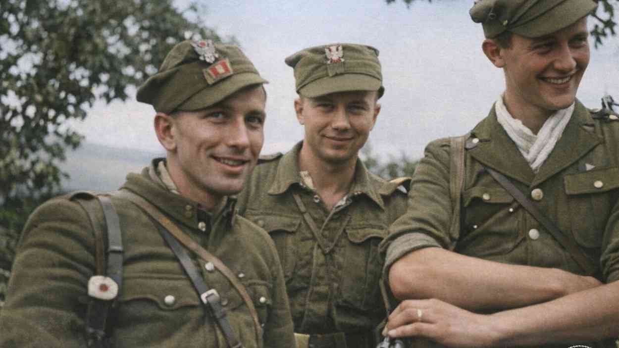 A colourised archival photo of three young soldiers in the uniforms of the 5th Vilnius Brigade, smiling and standing side by side outdoors.