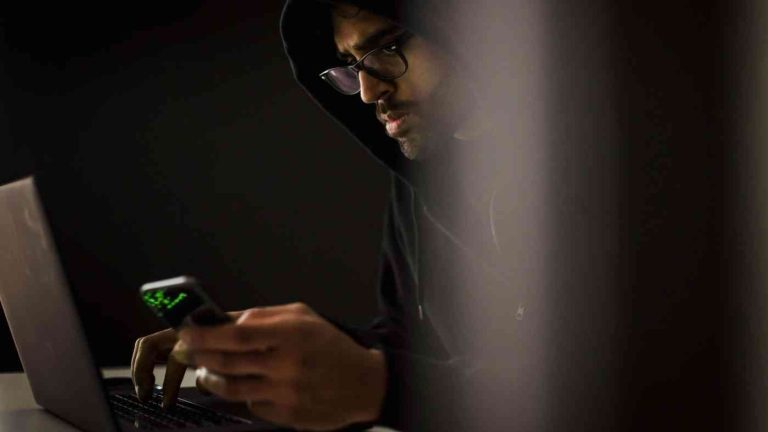 A hooded man sitting at a computer illustrates the relationship between the masculinity crisis and the manosphere online.
