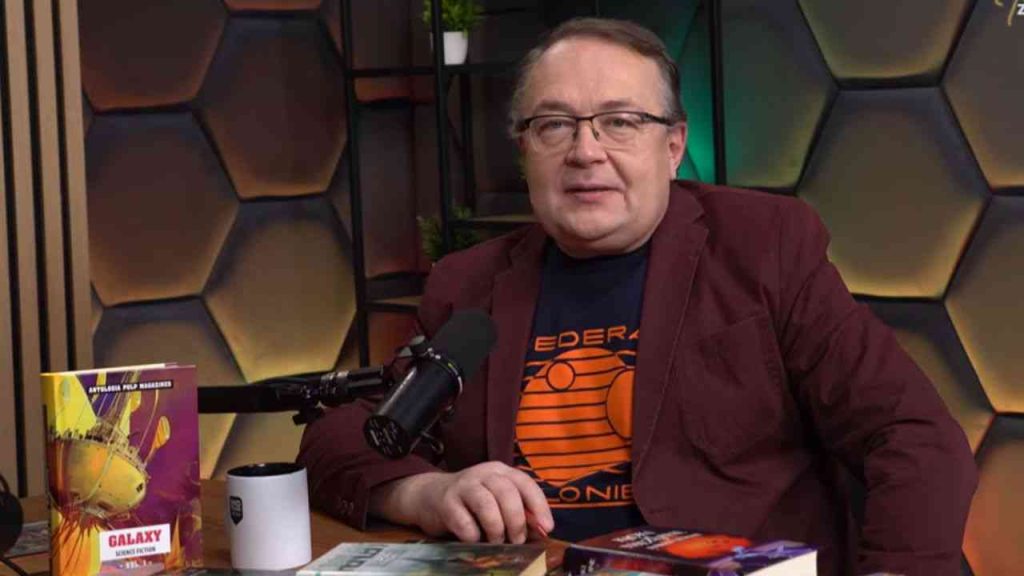 Piotr Gociek gives an interview. Medium close-up of a man’s face against a dark background, seated at a table.