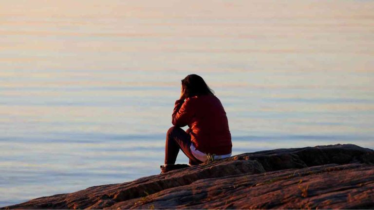 A woman sits with her back to the camera on a rock, gazing out over an endless expanse of water, wondering what happens after death.