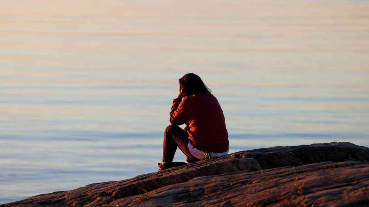 A woman sits with her back to the camera on a rock, gazing out over an endless expanse of water, wondering what happens after death.
